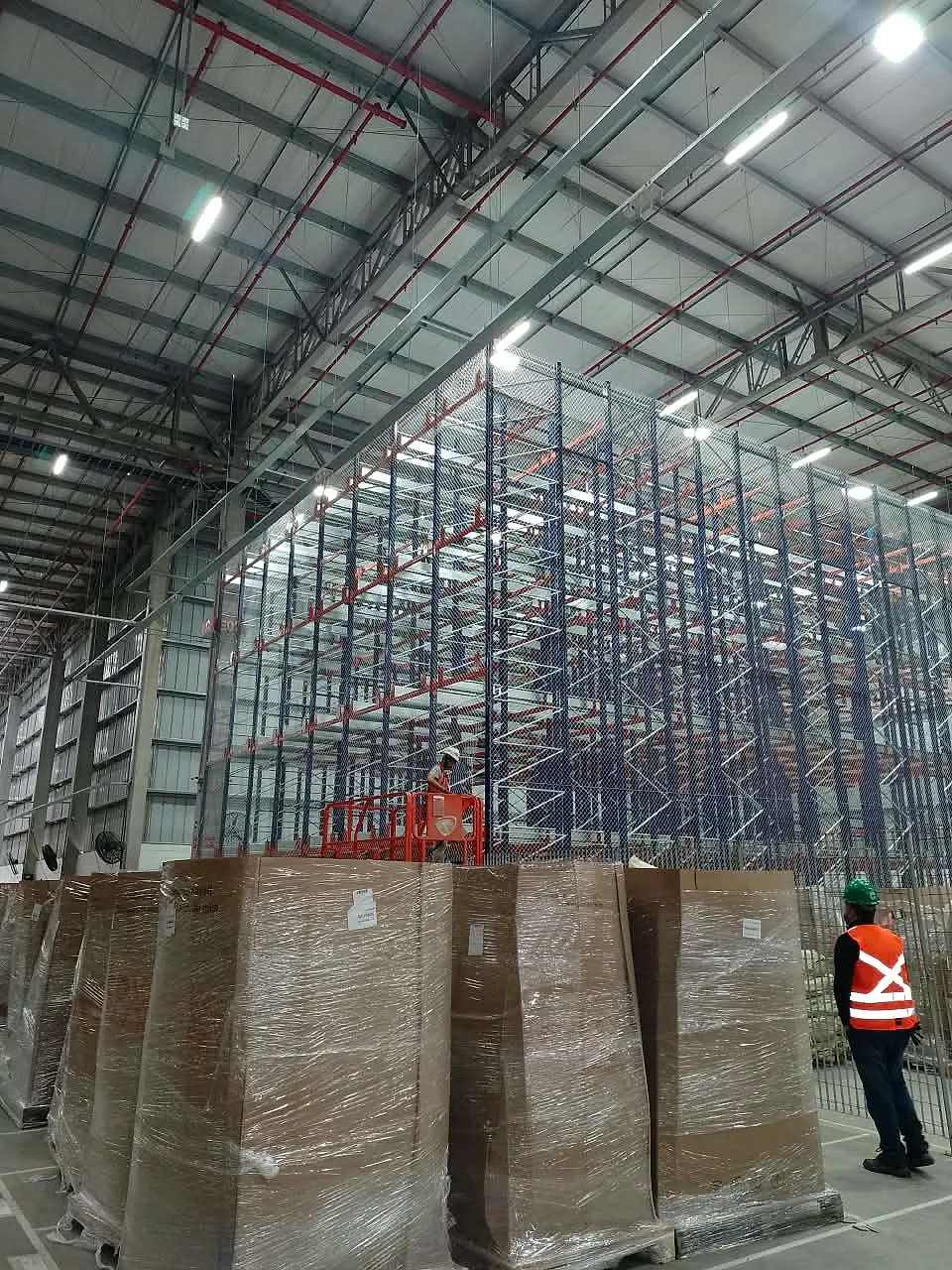 Técnico CARRÊ em plataforma elevatória tesoura instalando rede em porta-paletes de grande altura, com supervisor de segurança acompanhando do solo