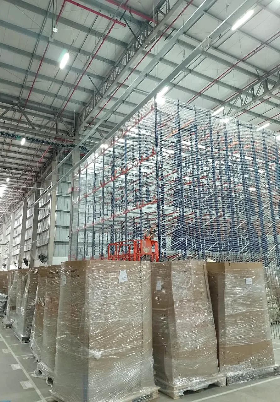 Vista geral de galpão logístico com bloco de porta-paletes de alta elevação recebendo rede de proteção CARRÊ e técnico em plataforma elevatória