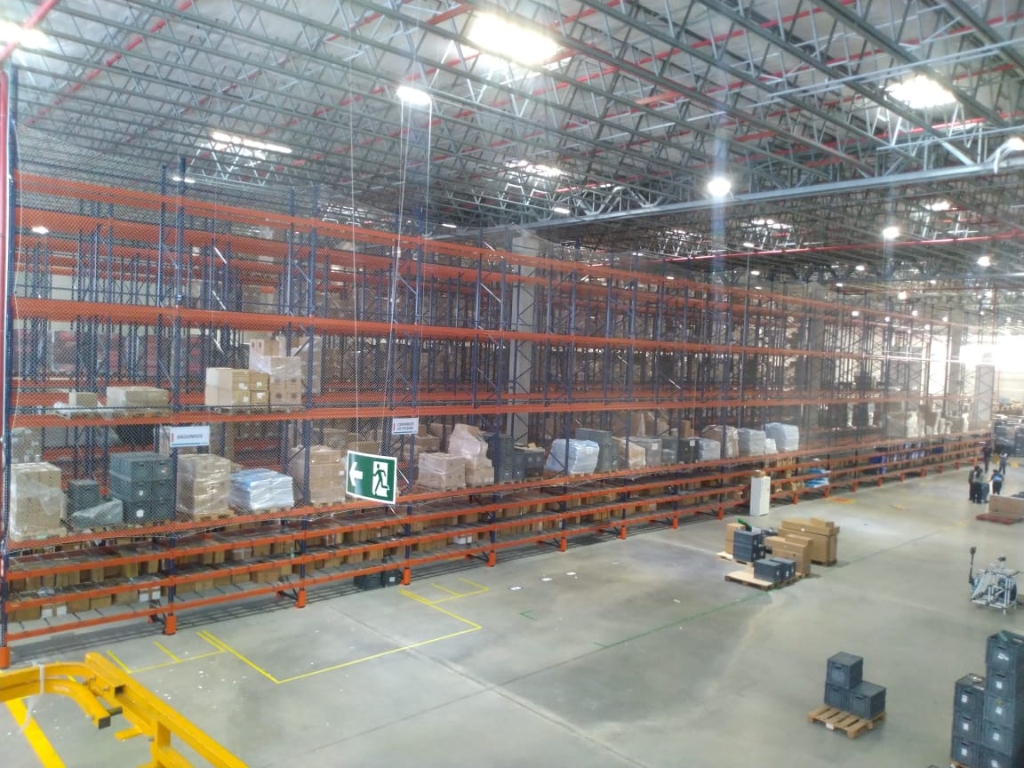 Vista panorâmica de galpão logístico de grande porte com linha extensa de porta-paletes totalmente protegida por redes de contenção CARRÊ acima da rota de fuga